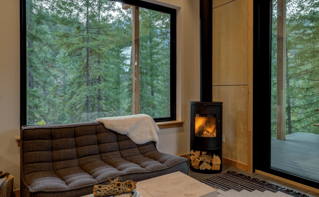 Interior view of living area shocasing a wood burning stove and couch under a large window with forest views
